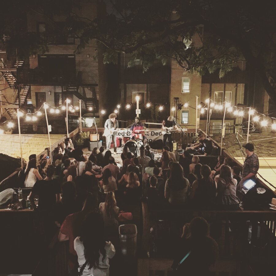 Seen from above at night, a packed backyard crowd under string lights watches Hunter-gatherer play on a small deck stage