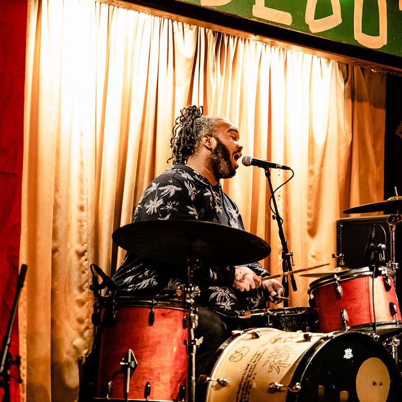 The drummer, locs swinging, singing into a mic behind a red drum kit in front of a gold curtain