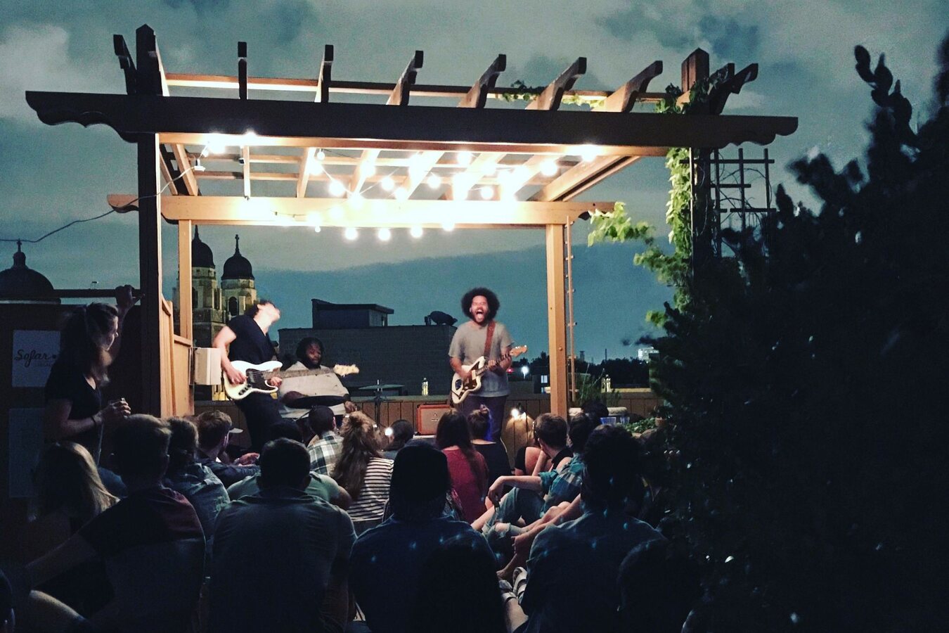 Hunter-gatherer playing a rooftop show at dusk under a pergola strung with bulbs, the singer shouting into the mic with his white offset guitar, church domes on the skyline and a seated crowd in front