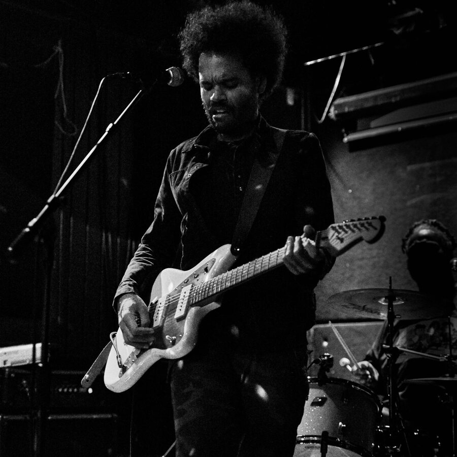 Black and white photo of the singer, eyes closed at the mic, playing a white offset guitar on a dark club stage